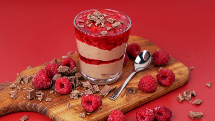 Glass med lagdelt dessert med sjokolademousse og bringebærkompott, omgitt av friske bringebær og sjokoladespon på treplate.