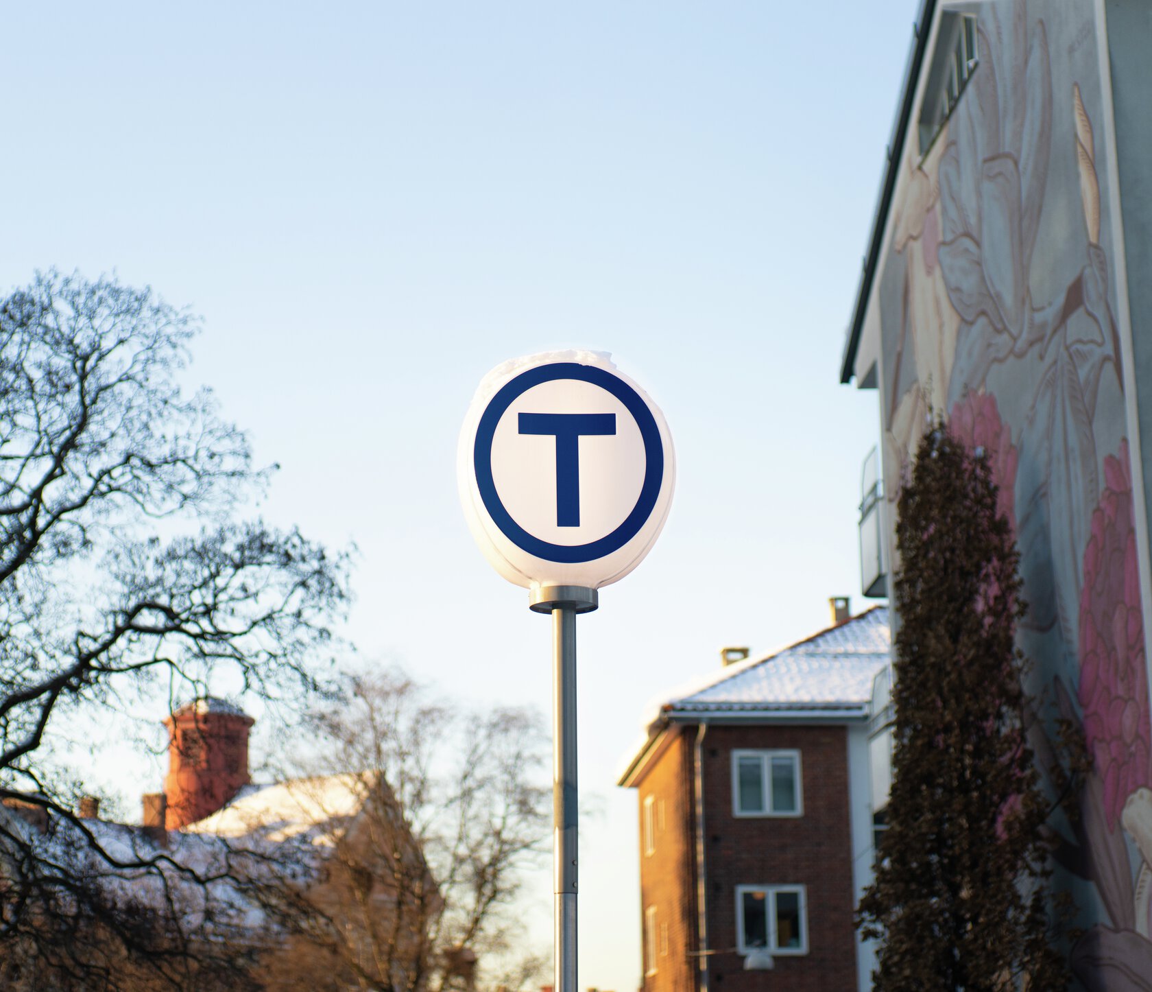 Foran trær og boligblokker står et rundt skilt med en stor   T  for T-bane. Foto