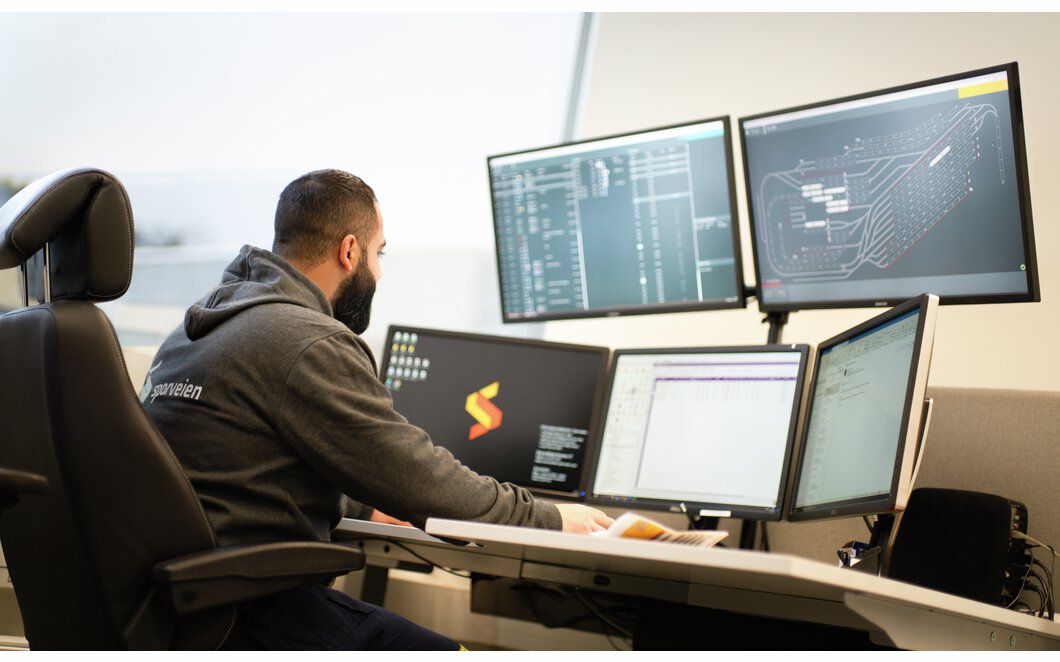 Mann med Sporveien-logo på genseren jobber foran 5 PC-skjermer. Foto.