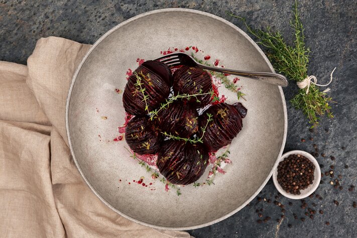 Hasselback-stekte rødbeter på en rustikk tallerken, garnert med frisk timian, servert med sorte pepperkorn i nærheten.
