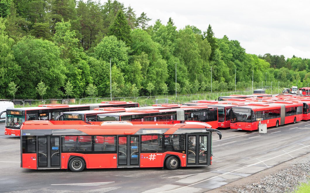 Mange busser står parkert foran en verkstedhall. Foto.