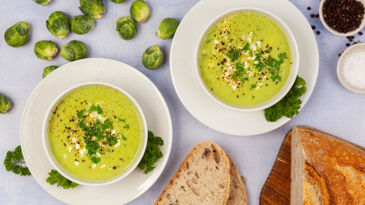 To boller med kremet rosenkålsuppe garnert med urter, feta og pepper, servert med sprøtt brødskiver.