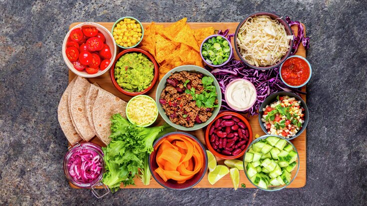 Oversikt over fargerik meksikansk matspredning: tortillas, guacamole, salsa, ost, grønnsaker, kjøtt og forskjellige pålegg på et trebrett.