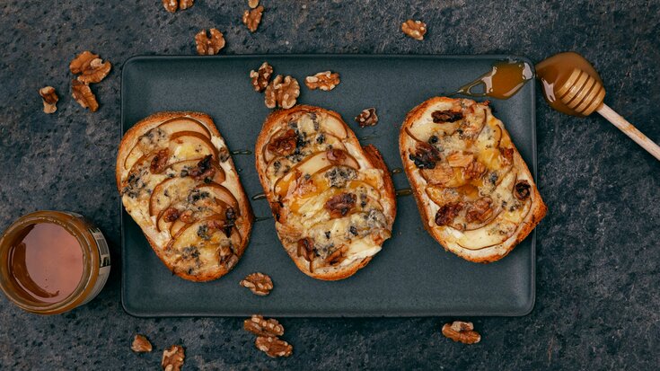 Tre åpne toasts toppet med smeltet brie, skivede pærer, valnøtter og honning på en mørk keramisk tallerken.