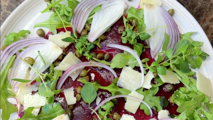 En livlig salat med ruccola, stekte rødbeter, rødløk, ostebiter og kapers på en hvit tallerken.