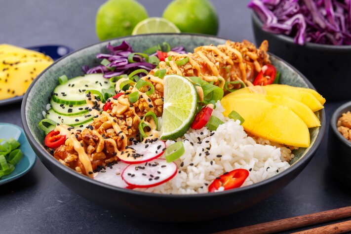 Fargerik poke bowl med ris, krydret kjøtt, mango, agurk, rødkål og topping. Omgitt av ingredienser og spisepinner.