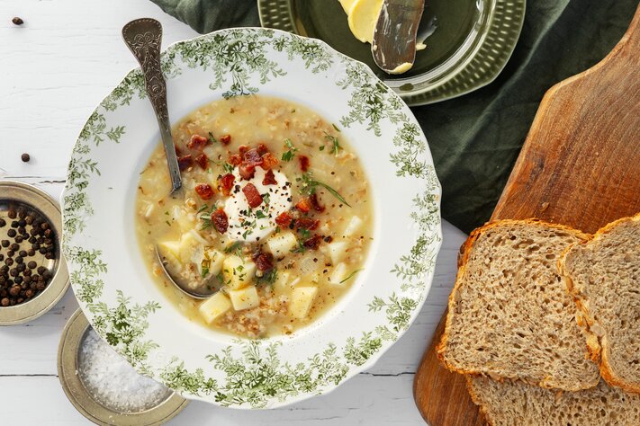 En bolle med kremet suppe toppet med krutonger og urter, servert med sesambrød på et trebrett.