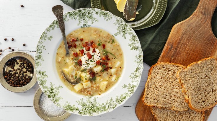 En bolle med kremet suppe toppet med krutonger og urter, servert med sesambrød på et trebrett.