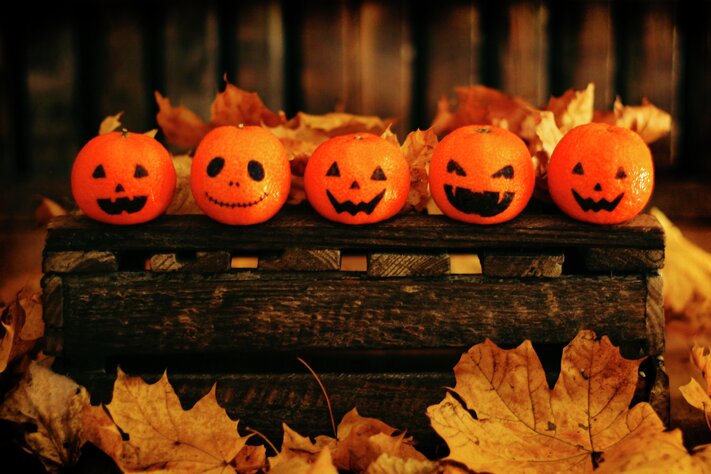 Fem appelsiner dekorert som jack-o'-lanterner sitter på en rustikk trekasse omgitt av høstløv i varmt lys.