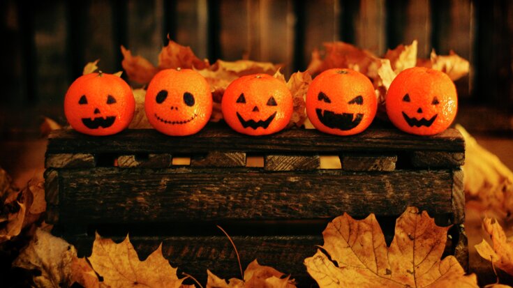 Fem appelsiner dekorert som jack-o'-lanterner sitter på en rustikk trekasse omgitt av høstløv i varmt lys.